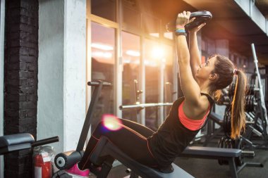Spor salonundaki egzersiz makinesinde oturan genç bayan ağırlık tabağını kaldırıyor..