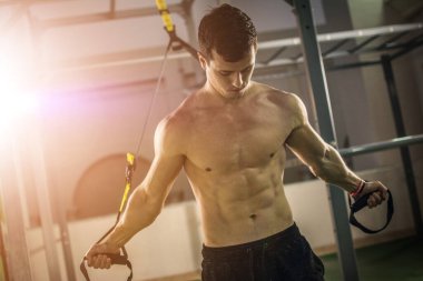 Spor salonunda kaslı, üstsüz, spor kemerleriyle antrenman yapan bir adam..