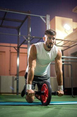 Yoğunlaşmış yakışıklı adam geceleri spor salonunda Ab Wheel ile egzersiz yapıyor..