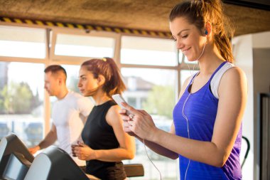 Spor giyim kuşağında gülümseyen kız telefonla müzik dinlerken spor salonunda koşu bandında yürüyor.