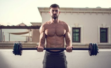 Gömleksiz kaslı adam dışarıda halterlerle egzersiz yapıyor..