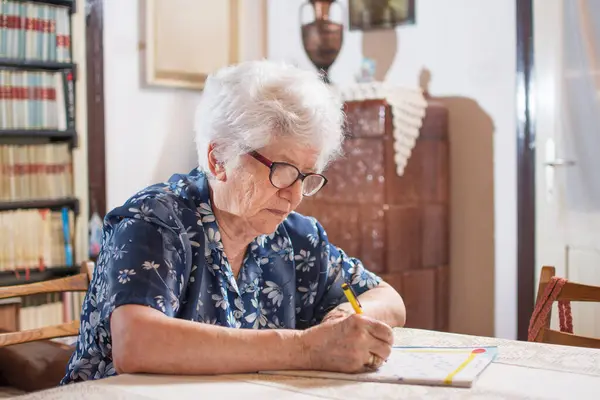 戴眼镜的老妇人坐在桌旁做填字游戏 — 图库照片