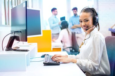 Bilgisayarda çalışan ve kameraya poz veren kulaklıklı IT servis operatörü kadın..