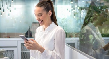 Resmi tişörtlü mutlu bir kadın. Saçları bağlanmış. Ofisteki akıllı telefonuna mesaj atıyor..