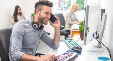 Yakışıklı iş adamı bilgisayar kullanıyor, cep telefonuyla konuşuyor ve ofiste çalışırken gülümsüyor.