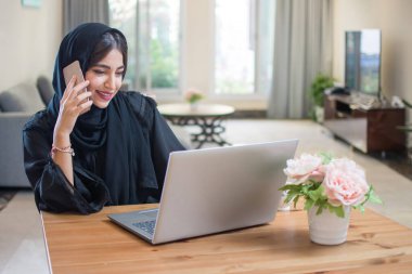 Güzel, genç, Müslüman, abaya giyen, cep telefonuyla konuşan ve evde dizüstü bilgisayarla çalışan bir kadın.