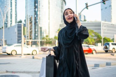 Güzel bir Arap kadını alışveriş torbaları taşıyor ve şehir caddesinde cep telefonuyla konuşuyor.