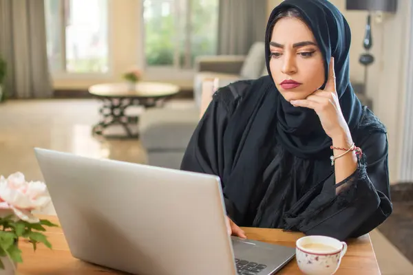 退屈な若いアラブの女性は 屋内で懸念される顔の表情で画面を見つめているラップトップでヒジャブを着ています — ストック写真