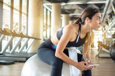 Fitness topunda oturan ve spor salonunda akıllı telefonlu müzik dinleyen aktif genç bir kadın. Rahatla ve spor antrenmanı konsepti