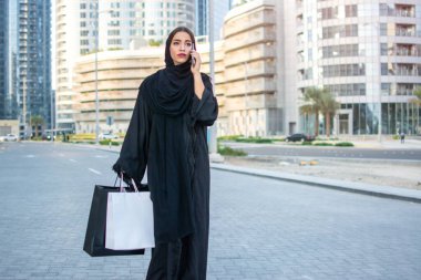 Güzel bir orta doğu Arap kadını Abaya giyer, alışveriş torbalarıyla sokakta yürür ve akıllı telefonuyla konuşur..