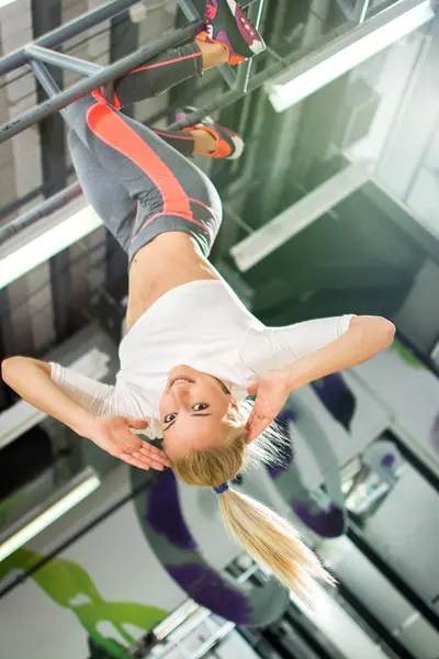 Young blonde sportswoman hanging upside down in a gym.