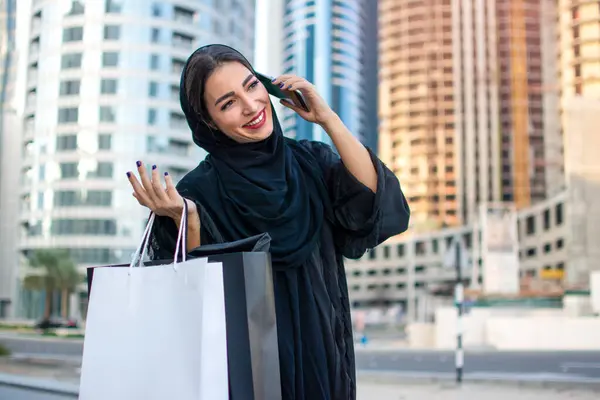 Arab woman in Abaya talking on mobile phone while holding shopping bags and walking on city street