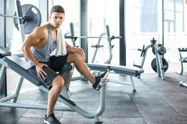 Spor salonunda boynunda havluyla egzersiz makinesinde dinlenen yakışıklı bir genç.