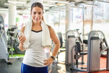 Omuzlarında havluyla gülümseyen sporcu kadın spor salonunda bir şişe su tutuyor..