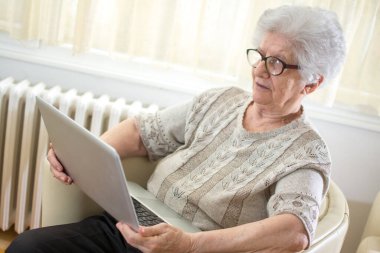 Gözlüklü, gri saçlı, dizüstü bilgisayar kullanan, evdeki koltukta oturan yaşlı kadın..