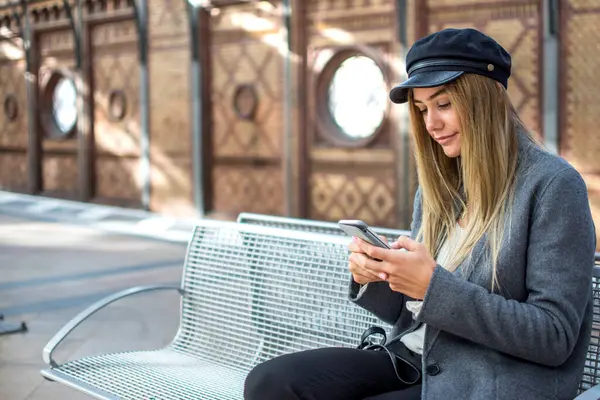 鉄道プラットフォームで電車を待っている間に電話で帽子をかぶっている美しい若い女性 — ストック写真