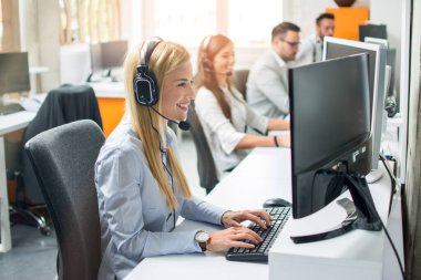 Kulaklıklı gülümseyen ajan kadın çağrı merkezinde ekibine eşlik eden bir müşteriye yardım ediyor.