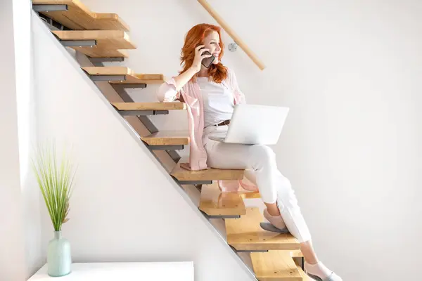 快乐的姜姑娘坐在家里的楼梯上 一边在膝上型电脑上工作 一边打电话 一边穿粉色衬衫 — 图库照片