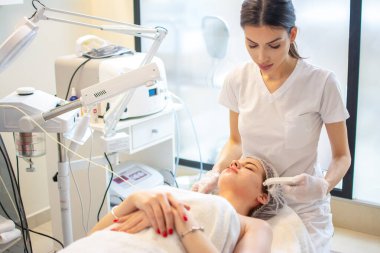 Güzellik uzmanı spa salonundaki genç bir kadının yüzüne modern aletlerle yüz bakımı uyguluyor.