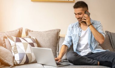 Genç, yakışıklı, gülümseyen adam kanepede oturup telefonla konuşurken bilgisayarını kullanıyor.