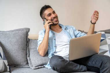 Genç, yakışıklı, gülümseyen bir adamın portresi. Telefonla konuşuyor ve evdeki kanepede otururken bilgisayarını kullanıyor.