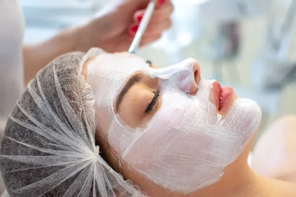 Face peeling mask, spa beauty treatment, skincare. Woman getting facial care by beautician at spa salon, side view, close-up.
