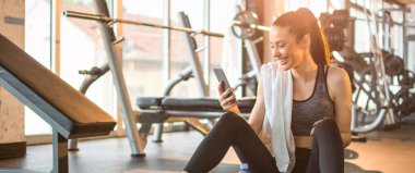 Spor salonunda spor yaptıktan sonra telefonu kullanan neşeli bir kadın. Spor salonunda telefon kullanan gülümseyen genç bir kadın..