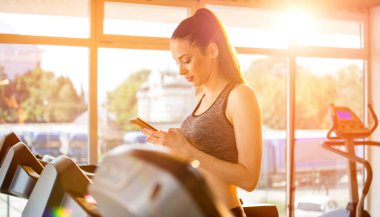Spor salonundaki koşu bandında yürürken telefonu kullanan genç bayan.