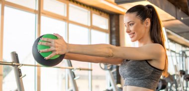 Genç, formda bir kadın spor salonunda diz egzersizi yapıyor. Güzel, gülümseyen bir kadın spor salonunda spor kıyafeti ve at kuyruğu giyerek sağlık topuyla egzersiz yapıyor..