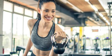Spor salonunda kettle Bell 'le antrenman yapan gülümseyen genç bir kadın. Spor giyim bölümündeki güzel fitness kızı sabahları çaydanlık çanlarıyla çalışıyor..