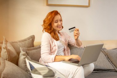 Genç mutlu kızıl kadın evde kanepede otururken alışveriş için dizüstü bilgisayar ve kredi kartı kullanıyor..