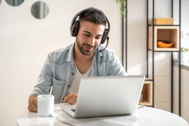 Evinden kahve içerken çağrı merkezinde çalışan dost canlısı genç bir adam. Kulaklık takmak ve dizüstü bilgisayar kullanmak..