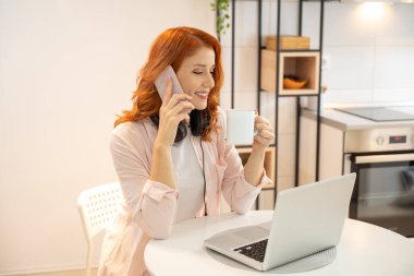 Genç gülümseyen kızıl saçlı çağrı merkezi ajanı telefonda, kulaklıkla konuşuyor ve dizüstü bilgisayar kullanıyor. Günlük giysiler giyip elinde bir fincan kahveyle..