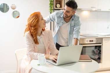 İki gülümseyen öğrenci laptoplarını birlikte çalışarak ve öğrenerek ya da video araması yaparak modern evdeki masanın önünde oturuyorlar. E-öğrenme.