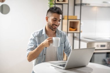 Evde dizüstü bilgisayarla çalışan gülümseyen genç adam. Günlük kıyafetler giyip, kahve içiyorlar..