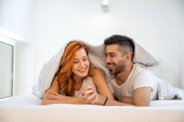 Beyaz battaniyeyle kaplanmış, el ele tutuşan ve evde yatakta uzanan genç bir çift. Beyaz tişört giyiyor..