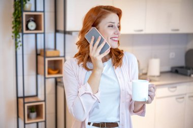 Genç, kızıl, güzel ve mutlu bir kızın portresi telefonda konuşurken çay içiyor ve sabahları mutfakta dikiliyor.
