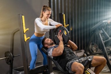 Genç ve kaslı bir adam spor salonunda genç ve çekici bir bayanın yardımıyla ağır tabağı kaldırıyor..