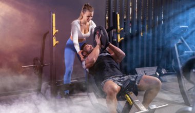 Genç ve kaslı bir adam spor salonunda genç ve çekici bir bayanın yardımıyla ağır tabağı kaldırıyor..