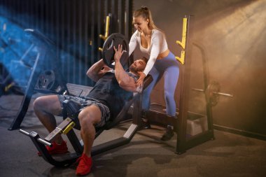 Genç ve kaslı bir adam spor salonunda genç ve çekici bir bayanın yardımıyla ağır tabağı kaldırıyor..