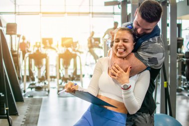 Kaslı zinde bir adam spor salonunda genç bir bayan antrenörü kol kaslarıyla boğuyor. Spor salonunda eğlenen arkadaşlar..