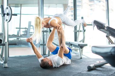Güçlü vücut geliştirici sırt üstü yatar ve spor salonunda sarışın bir fitness kızını bacaklarının üzerine kaldırır.