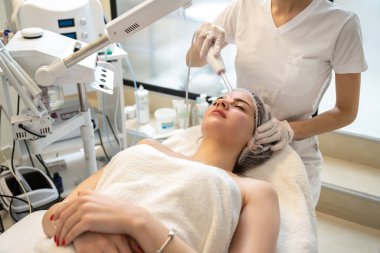 Güzellik uzmanı, güzellik salonunda bayan hastaların alnına özel bir aletle elektrikli Darsonval yüz masajı uyguluyor.