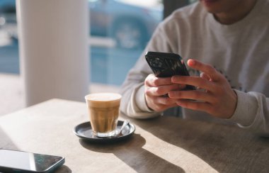 checking social media with smartphone drinking coffee in cafe against beautiful sunlight, having a relaxing moment