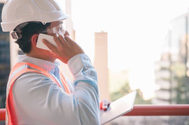 Dijital tablet ve akıllı telefon kullanan bir mühendis veya ustabaşı, erkek mimarın başlık takıp yeni binayı teftiş ederken, manüel işçisi, başlık ve gözlük giyerken görüntülendi.