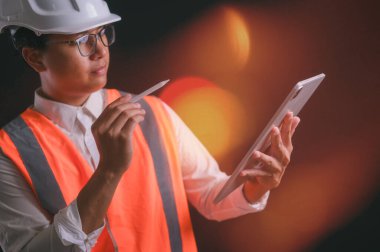 Gözlük takan mühendis, turuncu yelek ve dijital tablet tutan başlık.