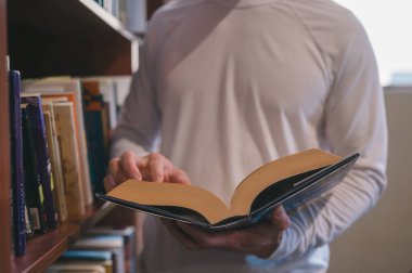 Kütüphanede kitap okuyan genç adam, yakın plan, üniversitede, üniversitede, üniversitede veya lisede ve edebiyat konseptinde kütüphanedeki kitaplıktan kitap tutan erkek elleri