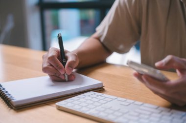 Masadaki Not Defterine Yazan Adam 'ın yakın plan görüntüsü, Bir kişi akıllı telefon ve klavye kullanırken bir kalemle bir not defterine yazar