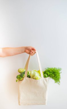 Çevre dostu bir yaşam tarzını ve plastik torbaların reddedilmesini sembolize eden, sebze ve yeşilliklerle dolu bir çantayı beyaz arka planda tutan bir el.