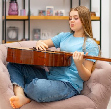 Genç kız koltukta oturur ve evde akustik gitar çalar. Huzurlu ve ilham verici müzikal ifade, hobi ve sıcak atmosferde kişisel gelişim anı